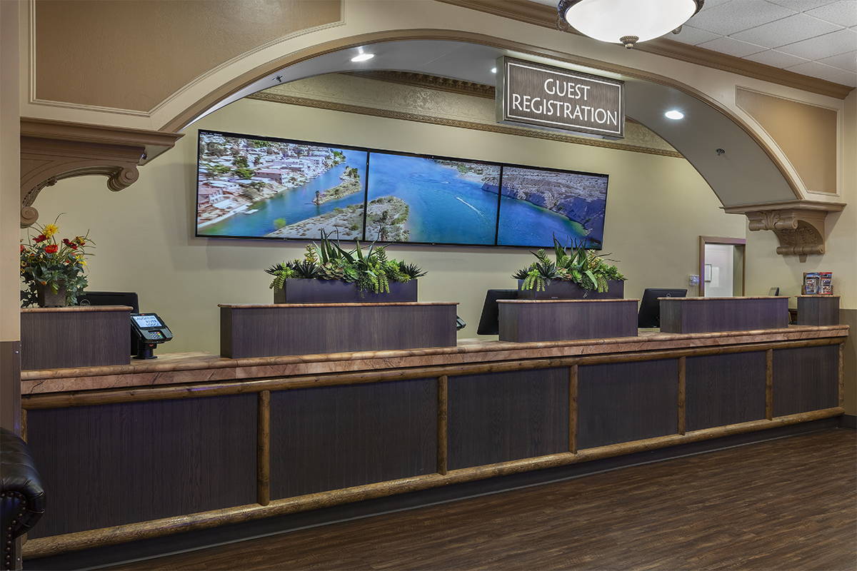 A hotel lobby reception desk with a wooden counter, plants, and screens on the wall.