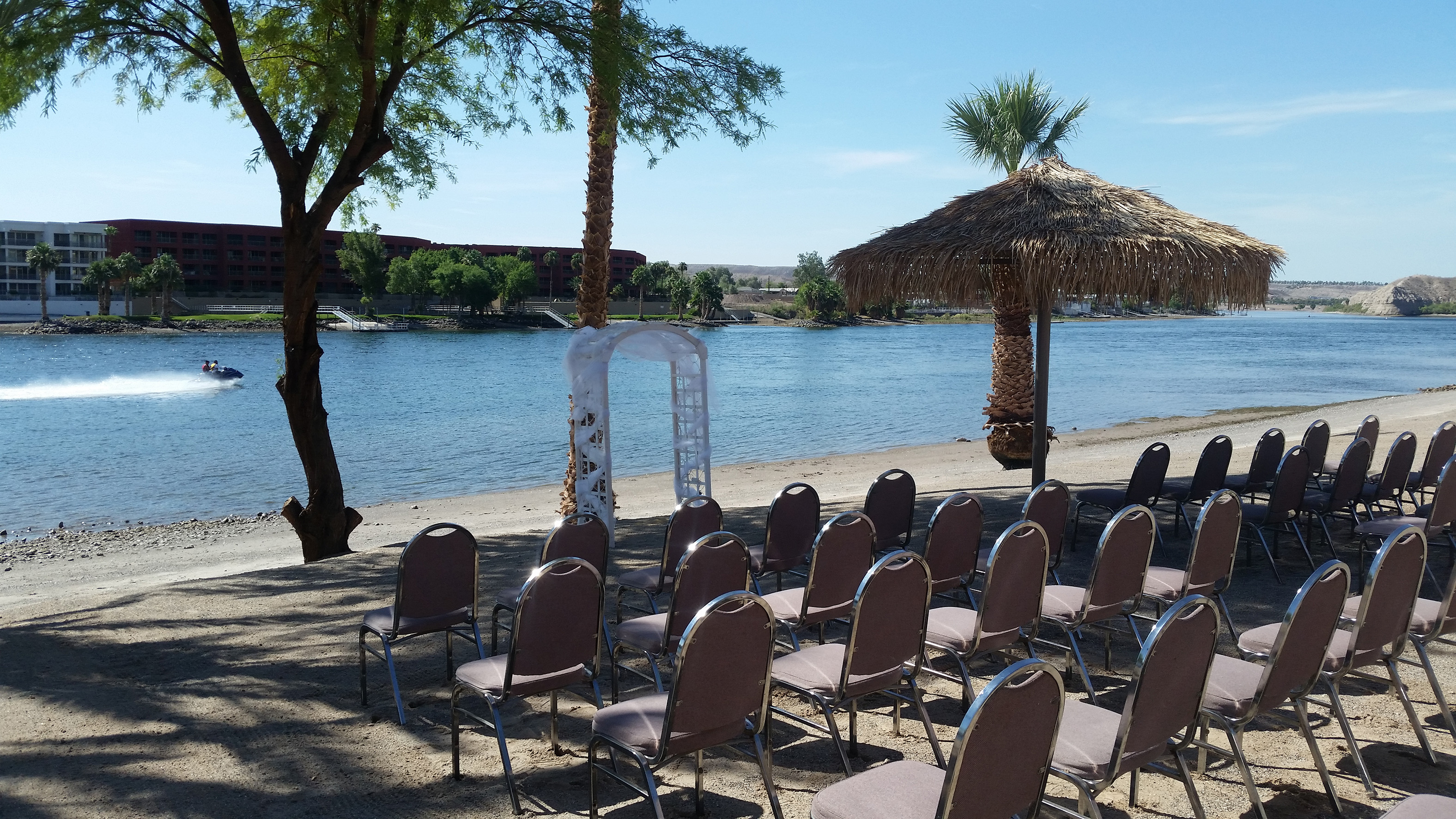 An outdoor event space with a river view, featuring rows of chairs, palm trees, a thatched umbrella, and a decorated archway.